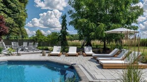 A swimming pool with lounge chairs and an umbrella, surrounded by trees and a wooden fence. - The Addison | Modern Farmhouse | Pool  and  Hot Tub (Prince Edward)
