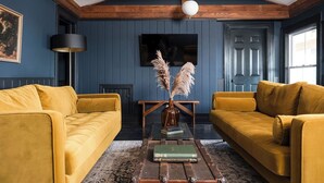 Une pièce avec deux canapés de couleur jaune moutarde, une table basse en bois sur laquelle sont posés des livres, un vase contenant des plumes d’herbe des pampas et un mur bleu foncé sur lequel est fixé un téléviseur.