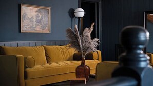 Une pièce avec un canapé jaune moutarde, un vase contenant des plumes d’herbe des pampas, un tableau encadré accroché au mur et un grand lampadaire.