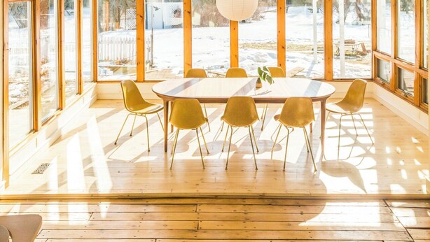 A spacious room with a large wooden ceiling, a dining area with a round table and six chairs, and large windows offering a view of a snowy outdoor area.