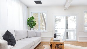A modern living room with a light-colored sofa, a wooden coffee table, a large plant, and a view through large windows to the outdoors. - The Hudson Guest House | Waterfront with Hot Tub (Prince Edward)