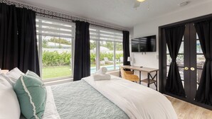 A hotel room with a large bed, a desk, a TV, and a view of a pool and garden. - SoFloasis - Home w/ Heated Pool, close to beaches (Lighthouse Point)