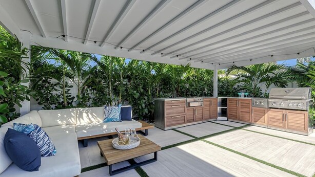 An outdoor patio with a white ceiling, a sofa with blue cushions, a wooden table with a basket, and a built-in barbecue station. - SoFloasis - Home w/ Heated Pool, close to beaches (Lighthouse Point)