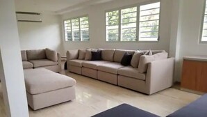 A spacious living room with a large beige sectional sofa, a smaller sofa, and a black ottoman. There are large windows providing natural light and a view of greenery outside. - Urban Paradise (San Juan)