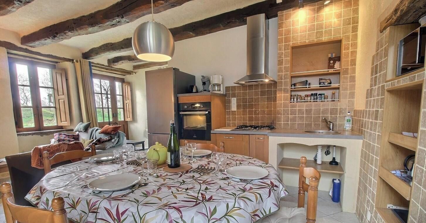 A dining area with a round table set for four, a kitchen with tiled backsplash, and a window with curtains.