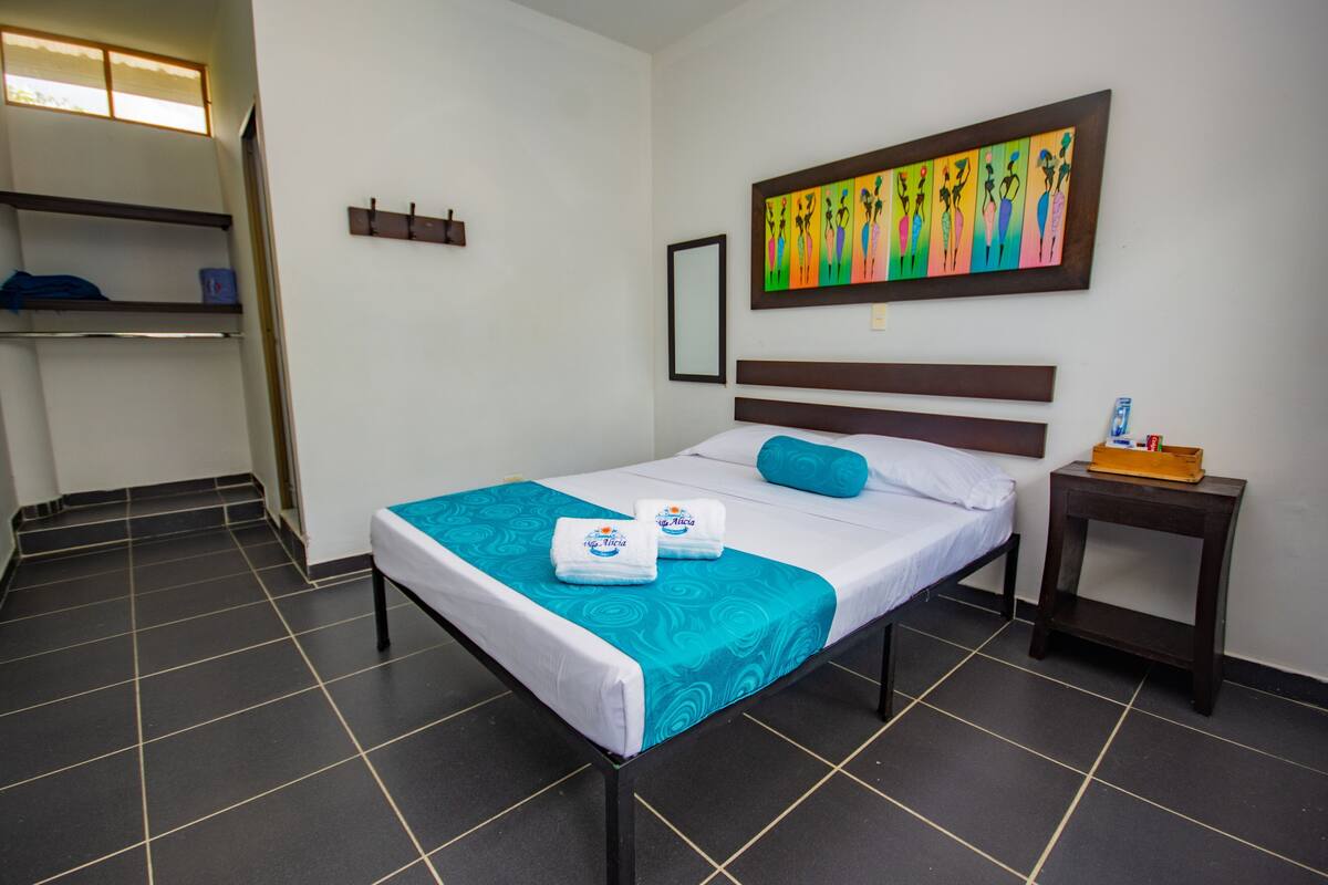 A hotel room with a bed, a nightstand, a colorful painting, a window, and a door.