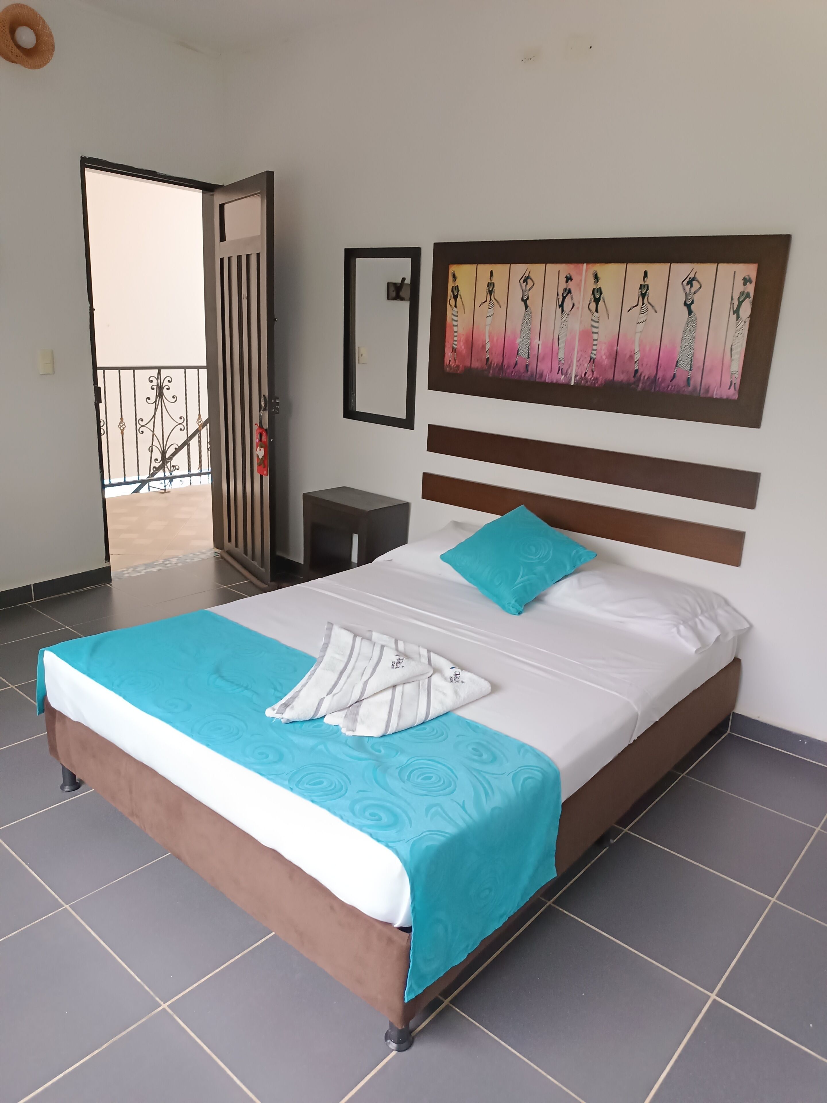 A neatly made bed with a blue and white bedspread, a single turquoise pillow, and a striped towel. A painting of women in dresses is on the wall above the bed.