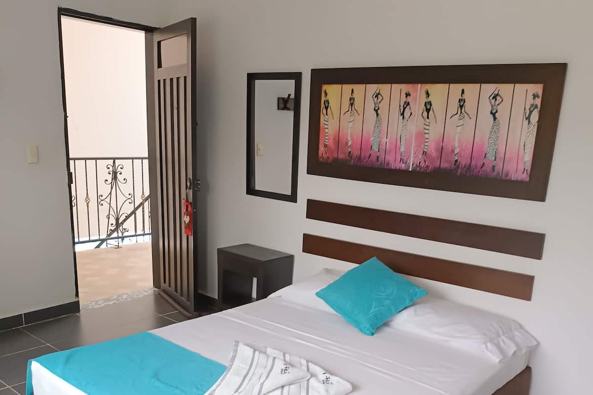 A neatly made bed with a blue and white bedspread, a single turquoise pillow, and a striped towel. A painting of women in dresses is on the wall above the bed.