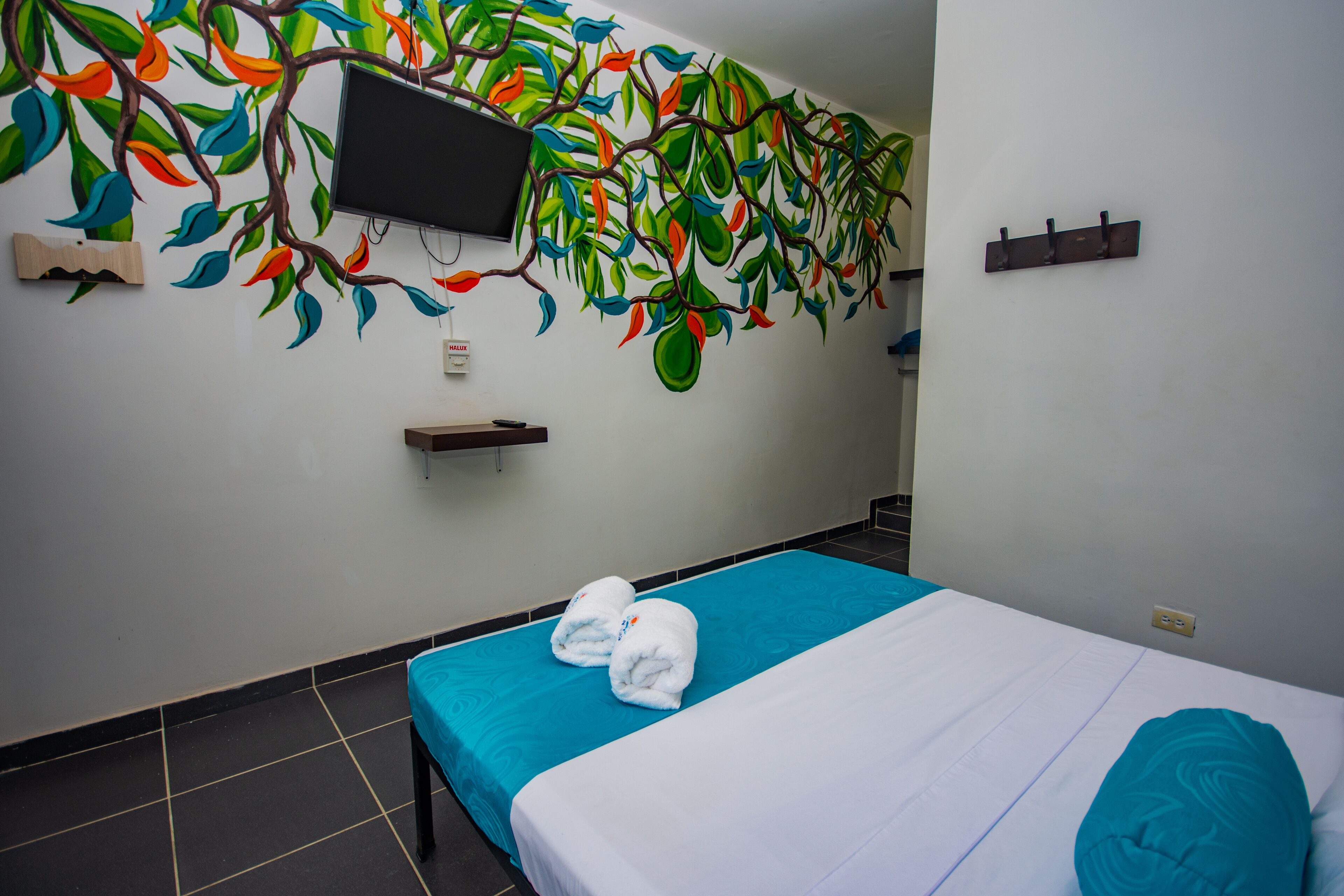 A hotel room with a bed, a blue bedspread, a teal pillow, a wall-mounted TV, and a colorful mural.