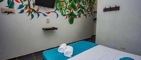 A hotel room with a bed, a blue bedspread, a teal pillow, a wall-mounted TV, and a colorful mural.
