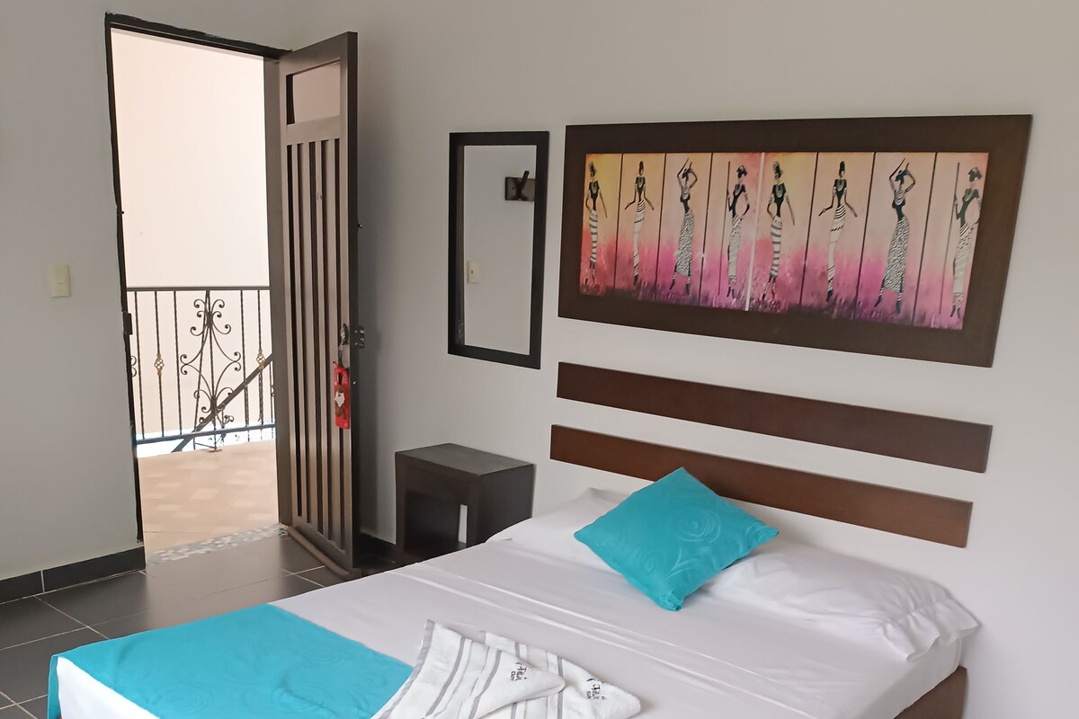 A neatly made bed with a blue and white bedspread, a single turquoise pillow, and a striped towel. A painting of women in dresses is on the wall above the bed.