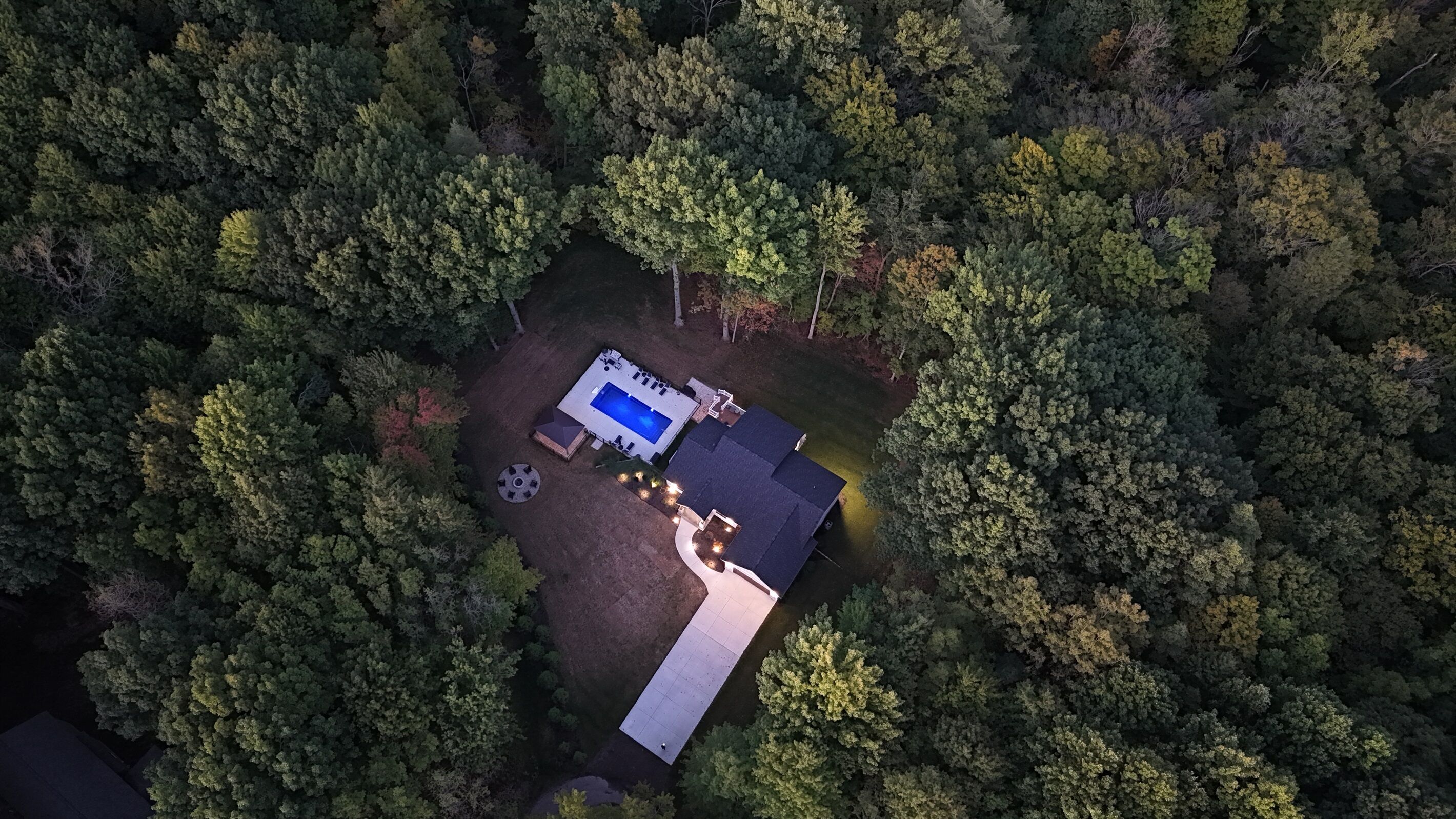 An aerial view of a property surrounded by dense forest, featuring a swimming pool, a covered patio, and a well-maintained lawn.