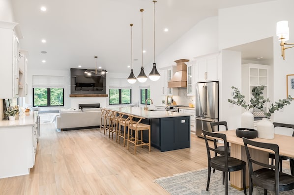 A modern kitchen with a large island, pendant lighting, and a dining area with a table and chairs. - Fall Retreat | Hot Tub, Games & Wine Country (South Haven)