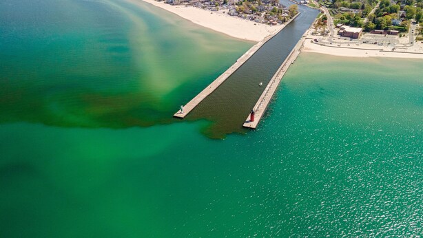 Een luchtbeeld van een kustgebied met een lange steiger die in het water reikt, omgeven door een woongebied met huizen en groen.