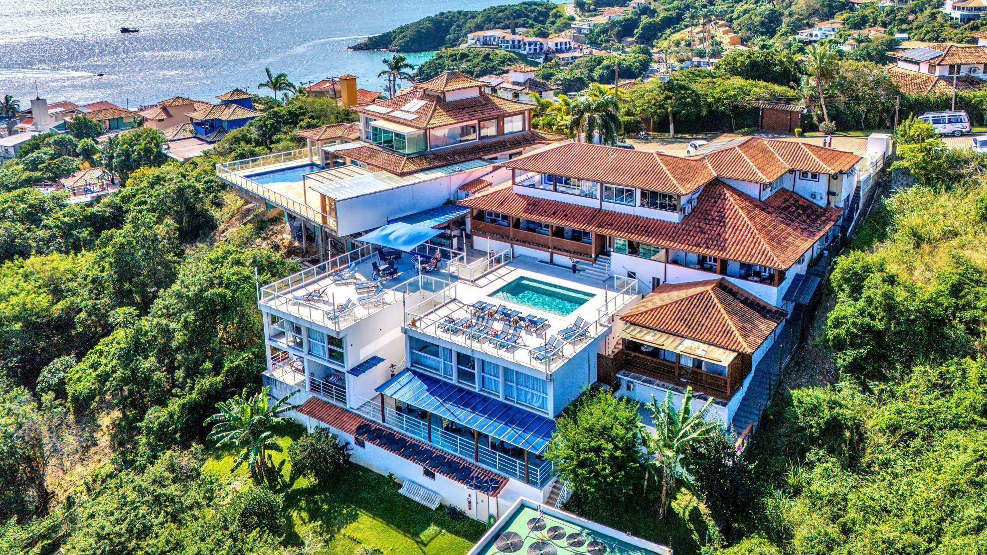 Edifício residencial com vários andares, piscina no terraço, rodeado por vegetação exuberante e vista para o litoral.