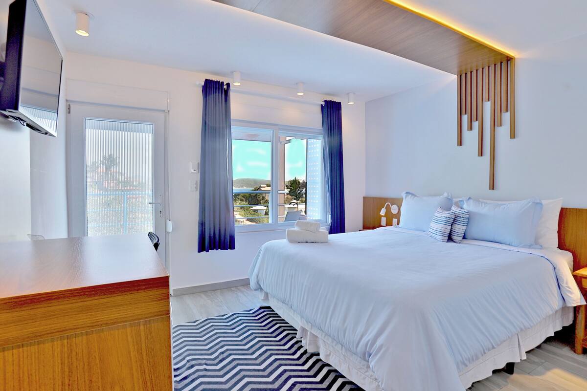 A modern hotel room with a large bed, a wooden desk, and a view of the outdoors through the window.