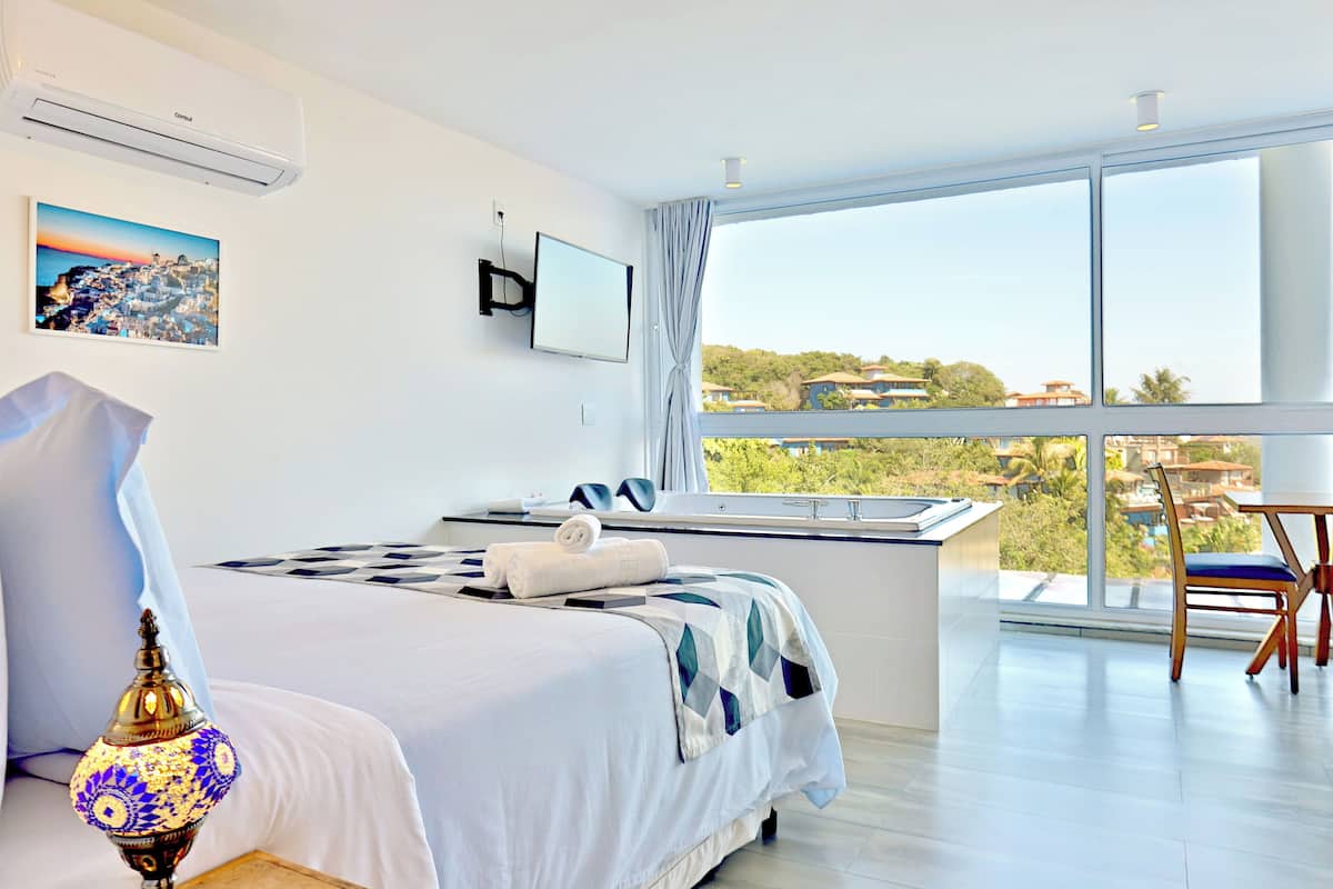 A modern hotel room with a large window, a bed with a patterned bedspread, a bedside table with a lamp, and a view of the landscape.