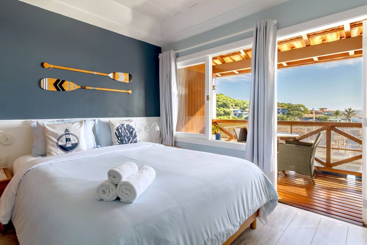 A bedroom with a bed, pillows, a wall-mounted paddleboard, and a balcony with a view.