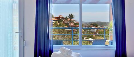 A hotel room with a bed, a window with blue curtains, and a view of a coastal town.