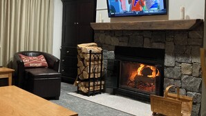A living room with a fireplace, a television mounted above it, a leather chair, and a table. - Okemo Ski in-Ski out @ Winterplace, 3 bdr, 2 ba Condo with Pool, Hot Tub, W/D (Ludlow)