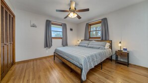 Een slaapkamer met een houten vloer, een bed met een bedovertrek met patroon, een nachtkastje met een lamp en een plafondventilator.