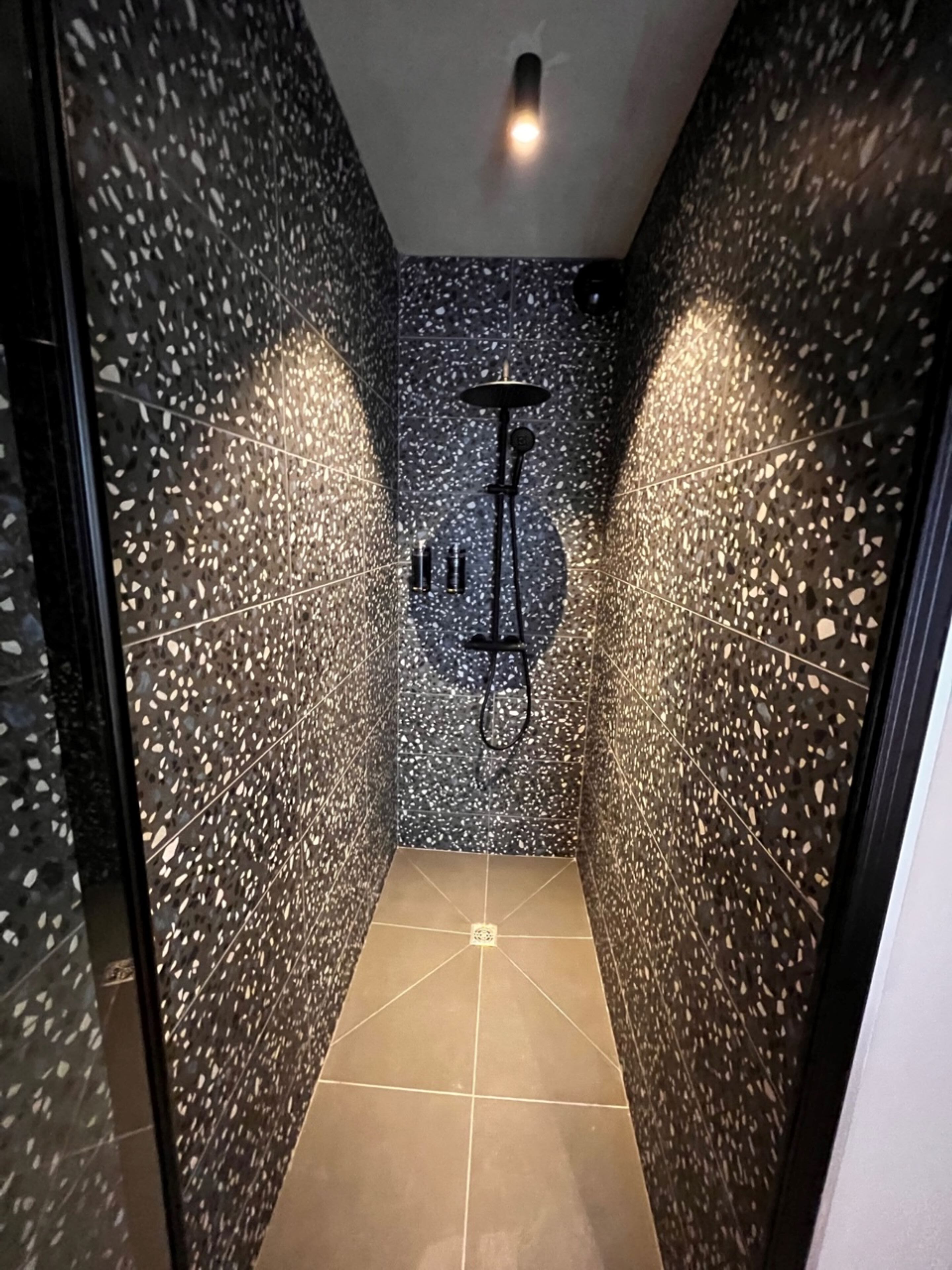 A narrow, tiled shower area with a showerhead and a small shelf with toiletries.