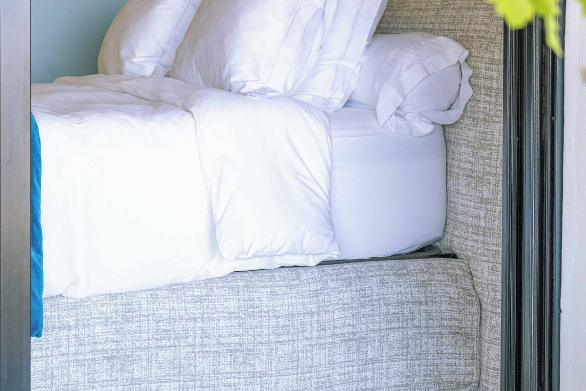 Una cama bien hecha con sábanas y almohadas blancas, un cabecero tapizado de gris, y vistas a una planta verde a través de una ventana.