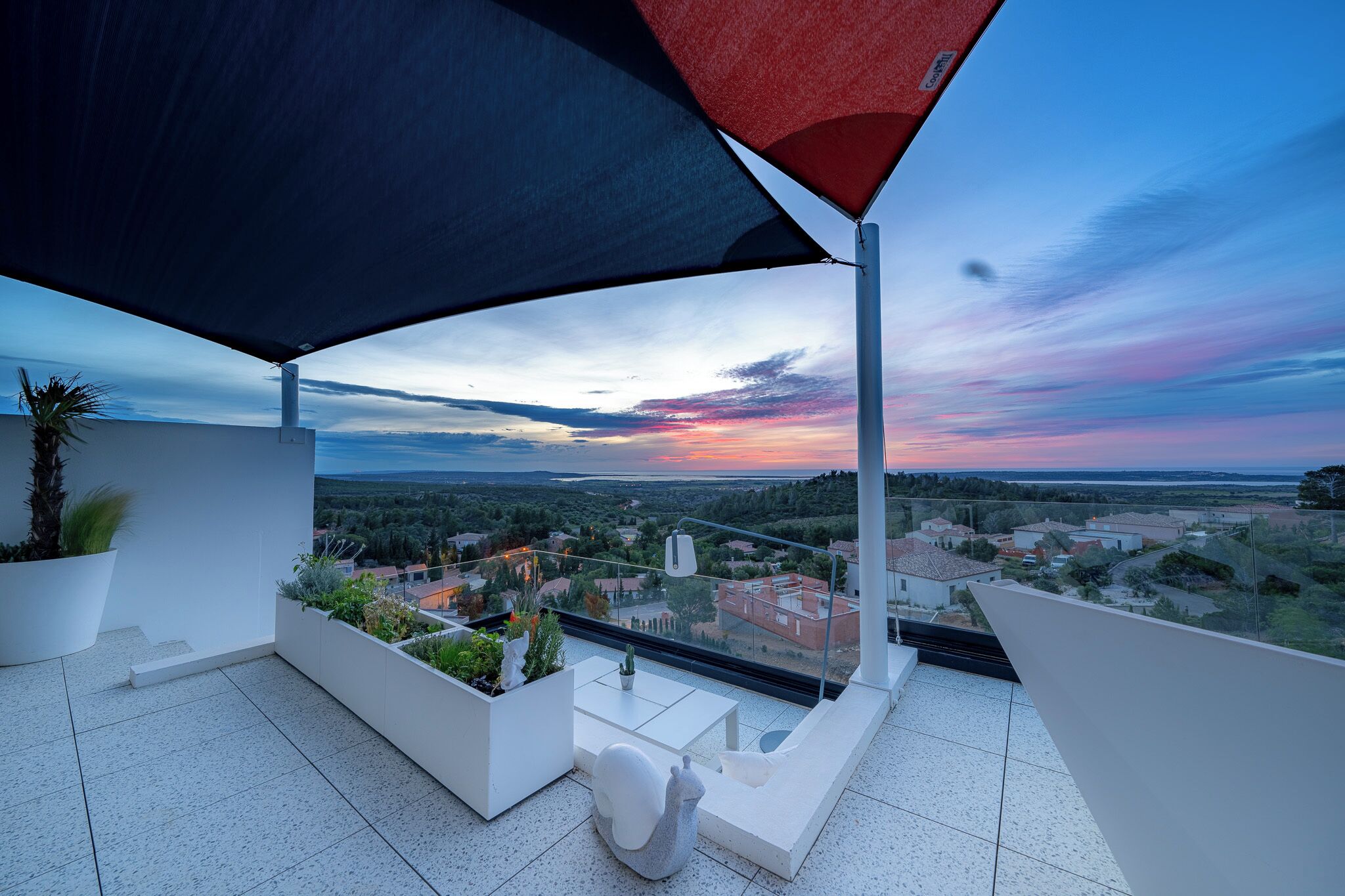 Un balcon offrant une vue sur un coucher de soleil se déclinant sur un paysage, des meubles d’extérieur blancs et un grand parasol pour créer de l’ombre.
