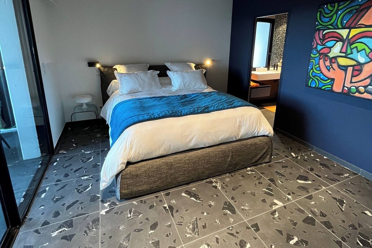 Un dormitorio moderno con una cama, un ventanal grande y un cuadro abstracto de colores en la pared.