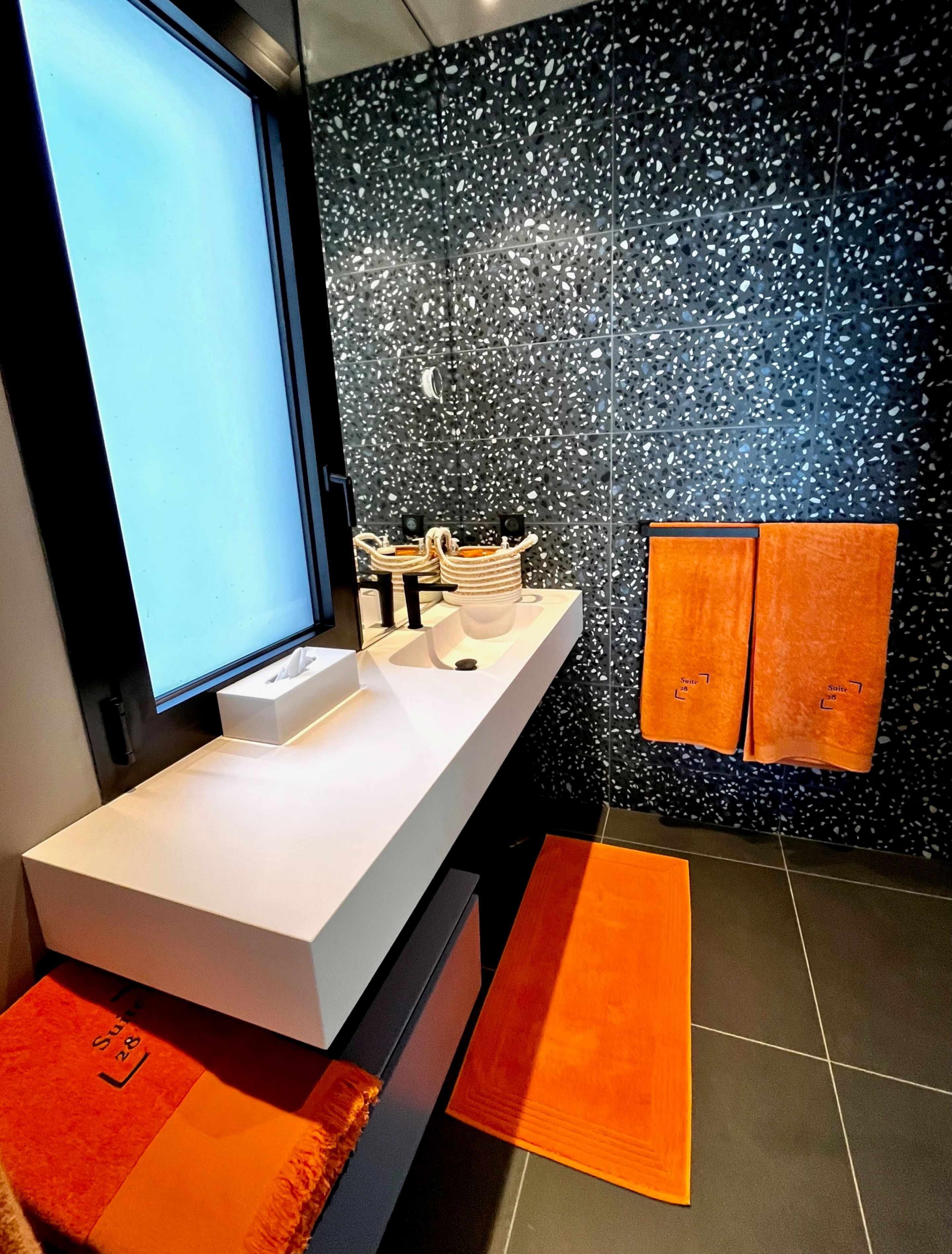 Une salle de bain moderne avec un meuble-lavabo blanc, un grand miroir et un mur à motifs noirs. Deux serviettes orange sont accrochées au mur et une boîte blanche est posée sur le meuble-lavabo.