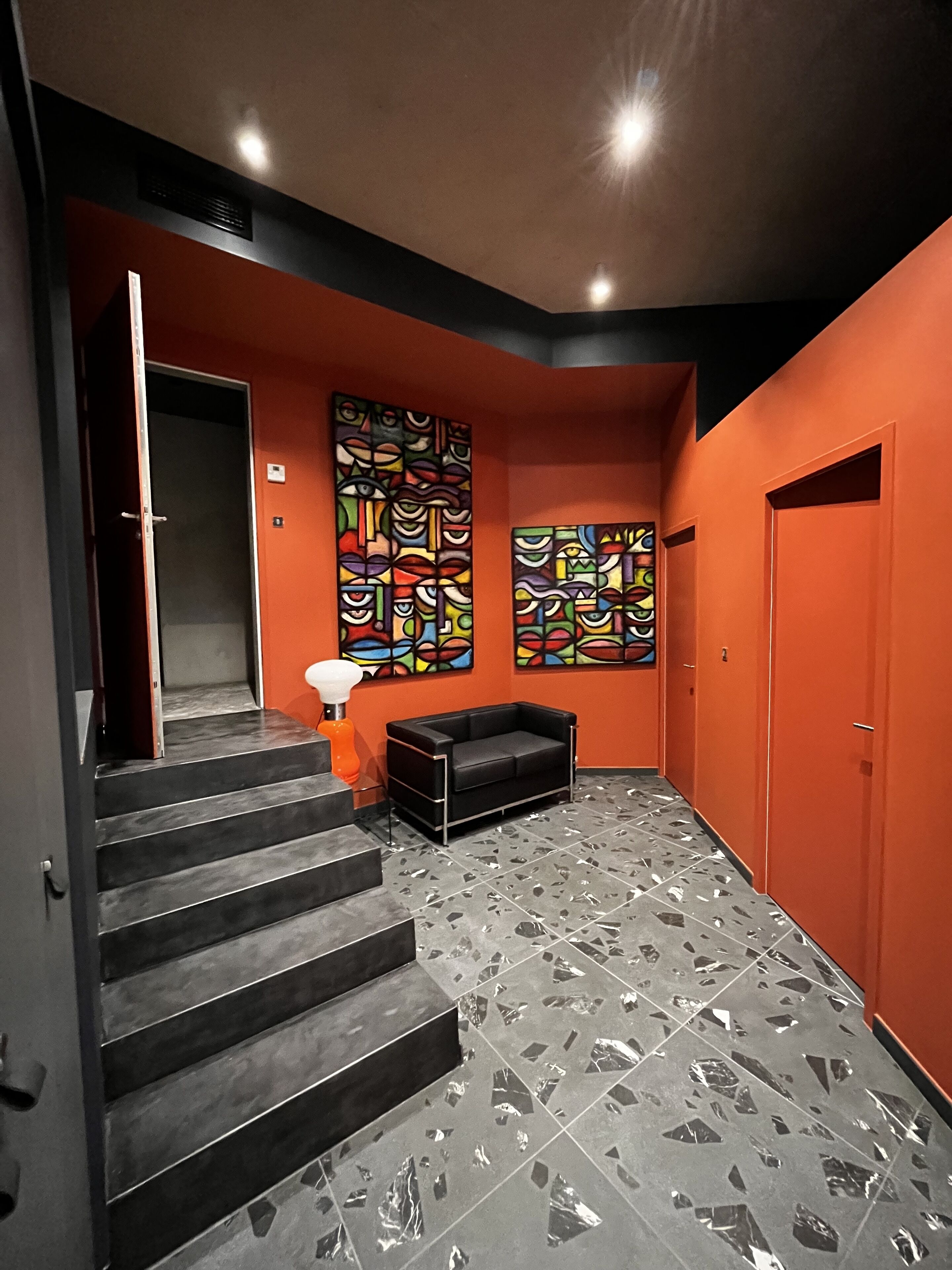 Een trap die leidt naar een kamer met oranje wanden, twee abstracte schilderijen, een zwarte bank en een betegelde vloer.