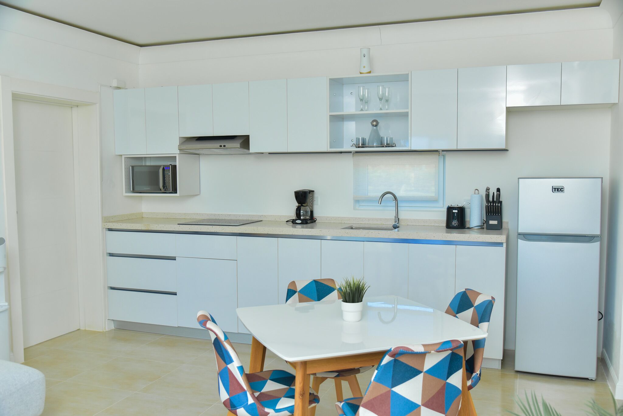 A modern kitchen with white cabinets, a microwave, a coffee maker, and a refrigerator. There is a dining table with chairs and a potted plant.