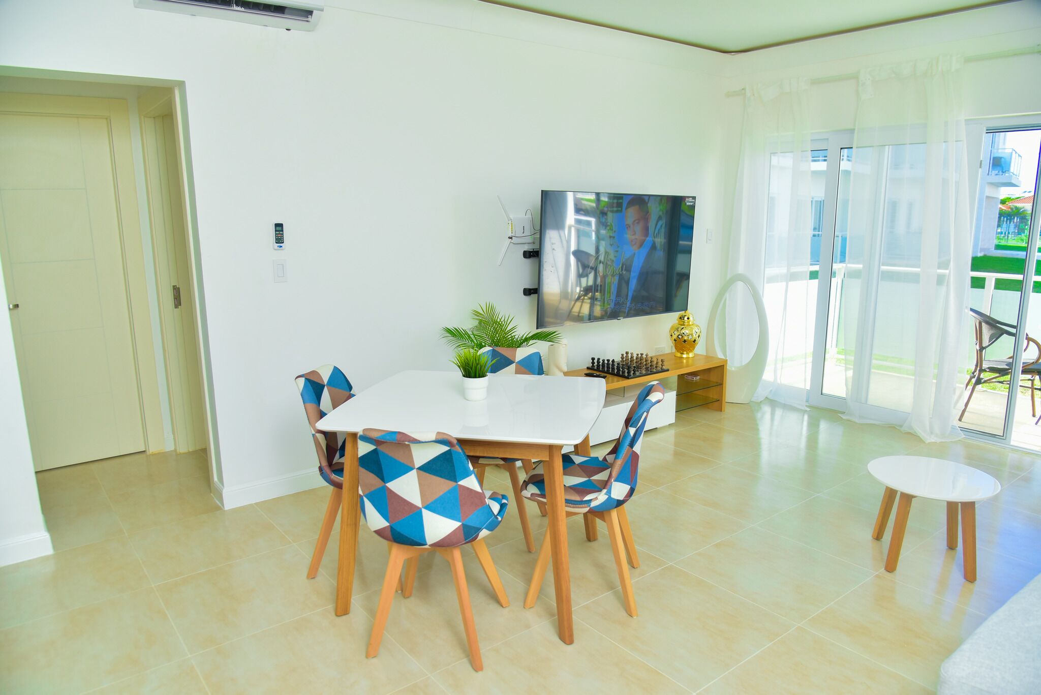 A dining area with a white table and chairs, a TV mounted on the wall, and a balcony with a view.