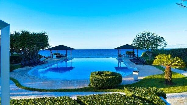 Piscina de un complejo turístico con vistas al mar, rodeada de tumbonas y un jardín cuidado.