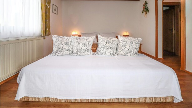 Una cama bien hecha con ropa de cama blanca y almohadas estampadas con motivos de hojas, dos lámparas de noche y suelo de madera.