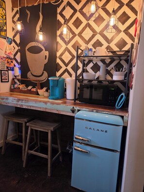 Une cuisine équipée d’un réfrigérateur Galanz d’époque, d’un four à micro-ondes et d’une machine à café posés sur le plan de travail. On y trouve également des tabourets et une étagère remplie d’ustensiles de cuisine.
