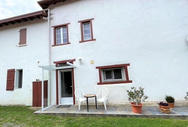 Un bâtiment blanc avec des fenêtres à encadrement rouge et une porte en verre. On y trouve deux chaises en plastique blanches et une petite table à l’extérieur.