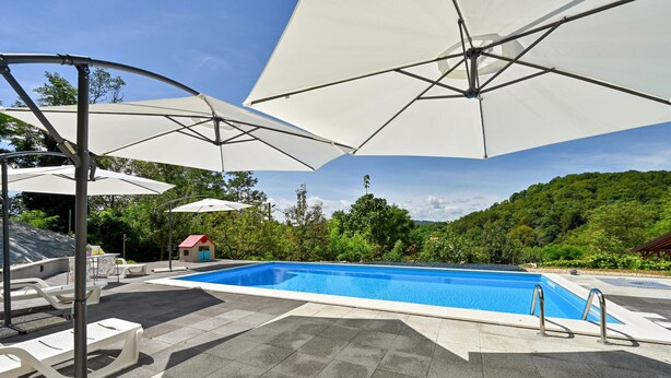Piscina con sombrillas blancas y sillones de descanso, rodeada de un jardín con árboles y vistas a una colina.