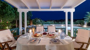 Un área de comedor al aire libre con una pérgola cubierta, sillas blancas y una mesa para dos, con platos, cubiertos y velas.