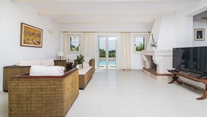 Amplia sala de estar con un televisor grande, mobiliario de mimbre y vista a una piscina a través de puertas de cristal.