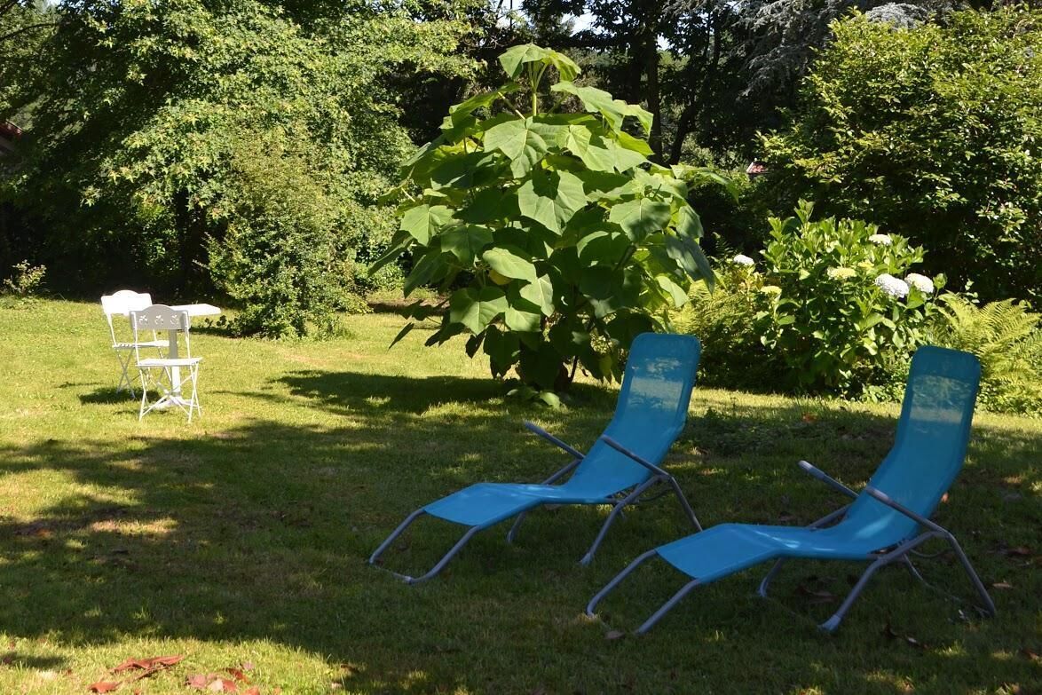Dos sillones azules en un área verde al aire libre, con un árbol grande y una mesa blanca al fondo.
