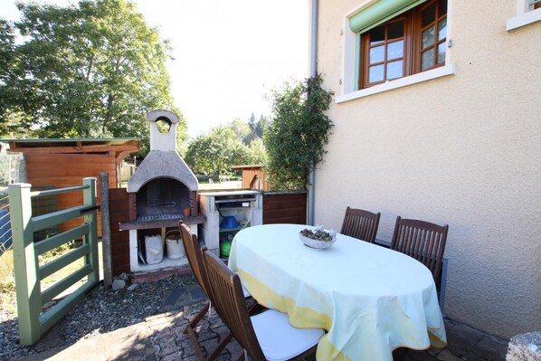 Eine Terrasse im Freien mit einem gemauerten Pizzaofen, einem Tisch mit weißer Tischdecke und Holzstühlen.