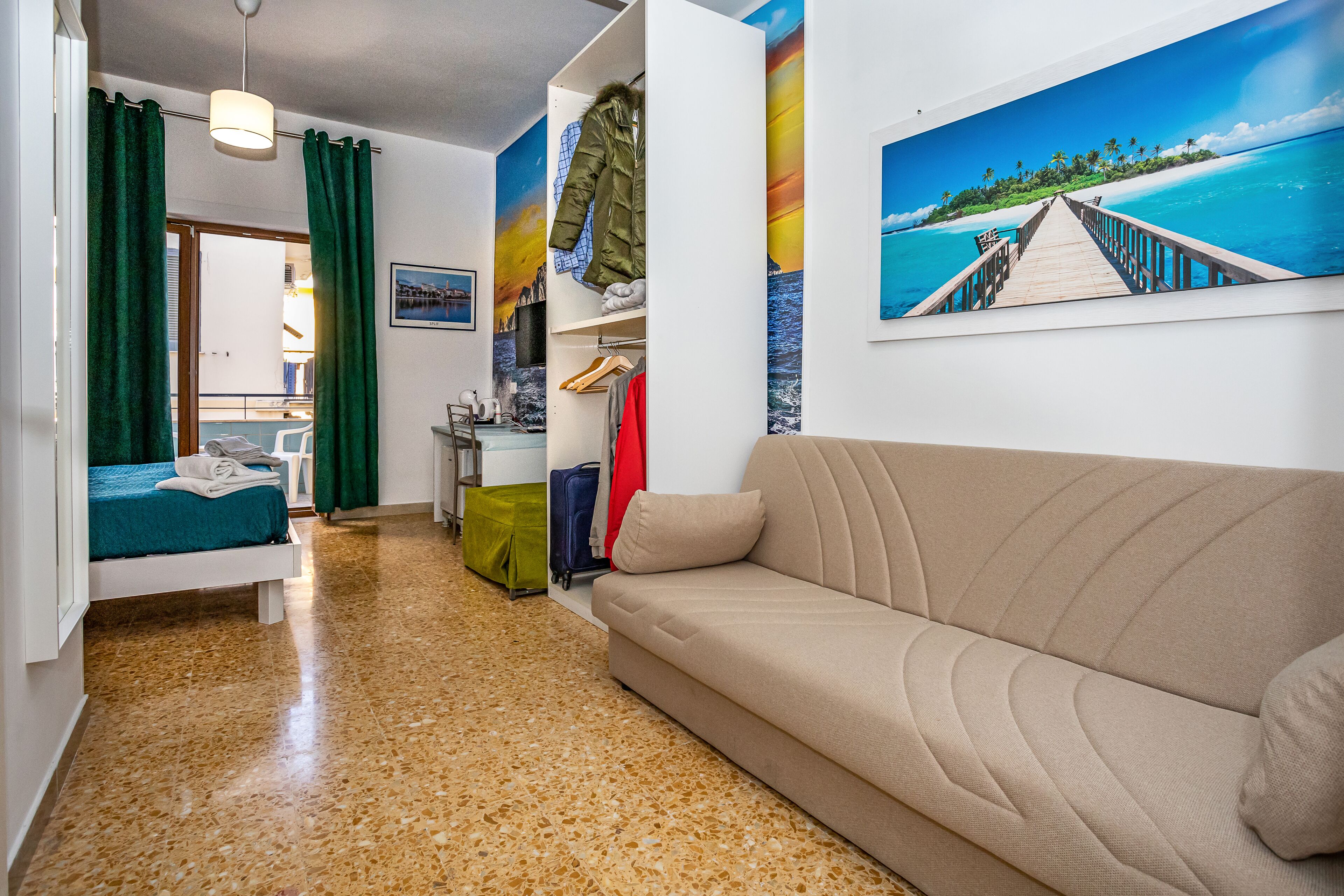 Habitació amb un sofà, un llit amb cortines verdes, un quadre de platja i un paviment de fusta.