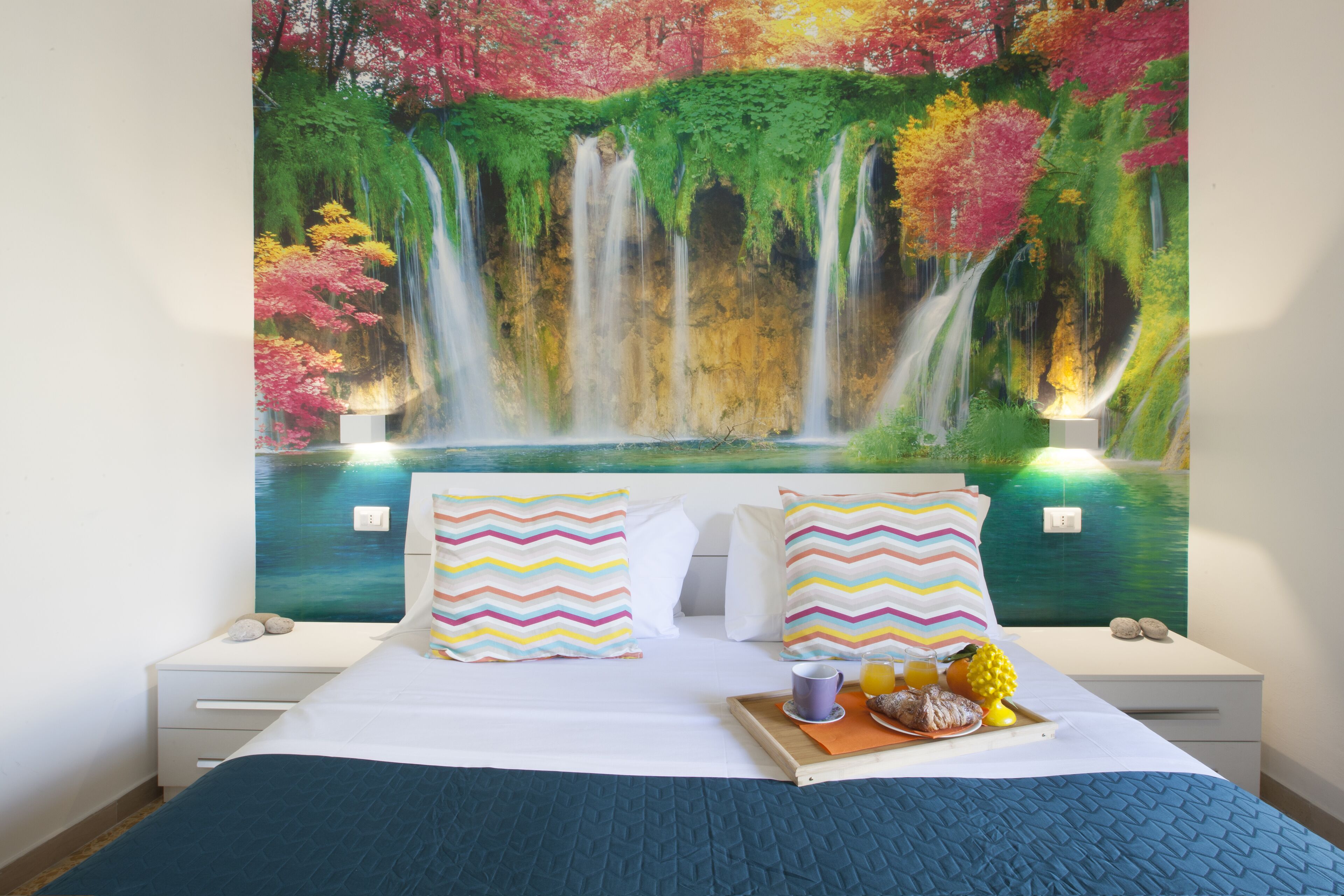 Un dormitorio con cama, almohadas, una mesita de noche, una bandeja de desayuno y un mural en la pared con una cascada.