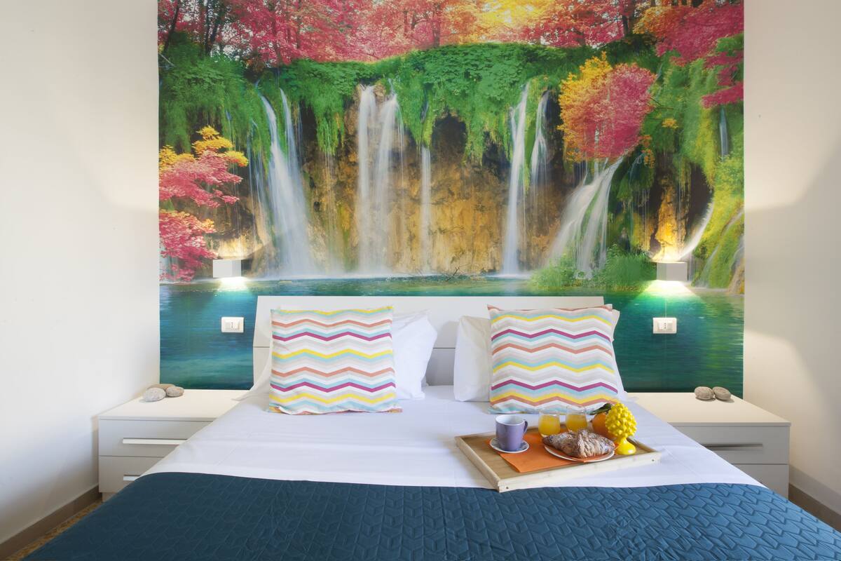 Une chambre à coucher avec un lit, des oreillers, un plateau avec des fruits et une tasse, et une peinture murale représentant une cascade.