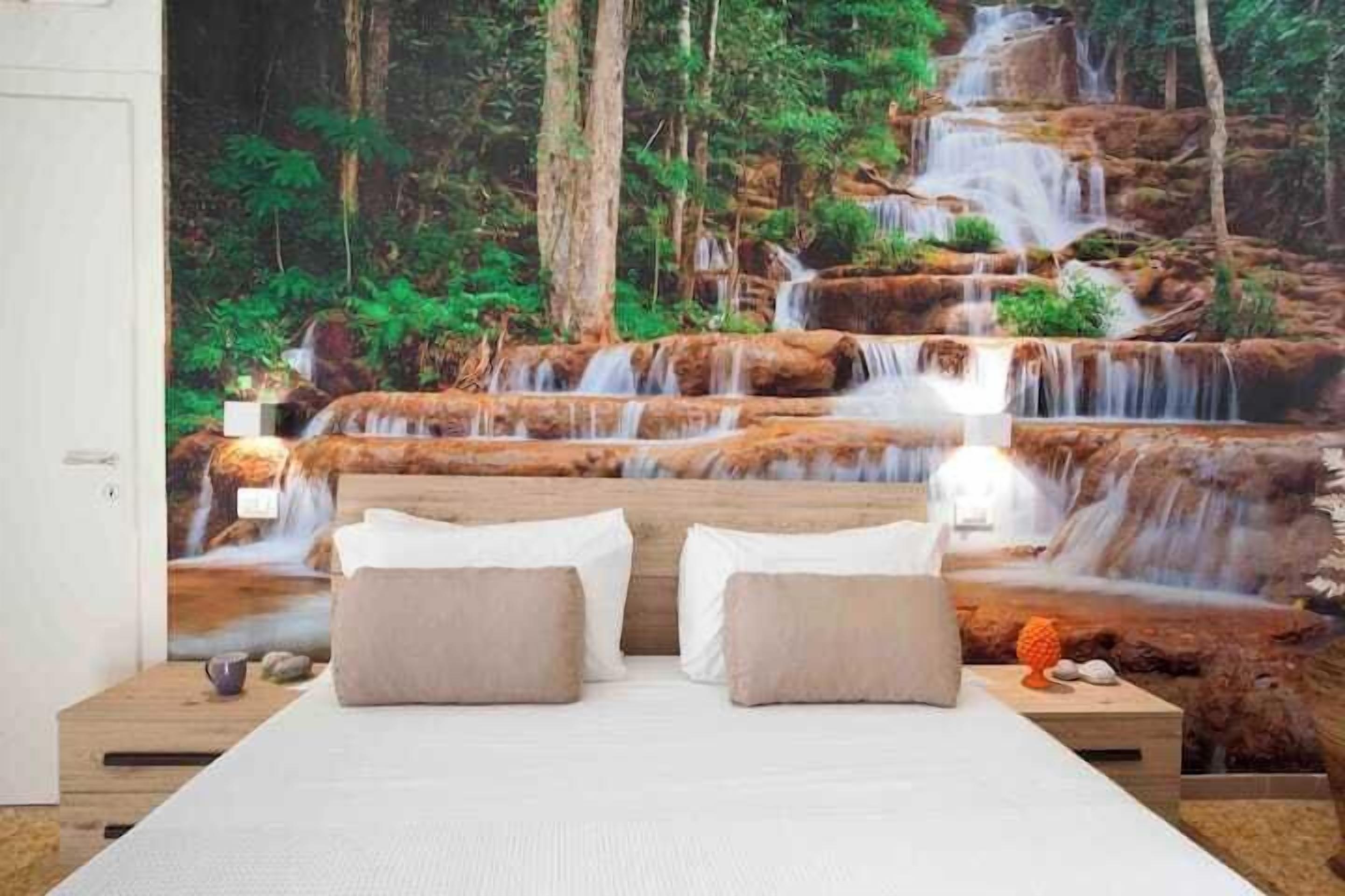 Quarto com cama, almofadas, mesa de cabeceira e um mural na parede com uma cachoeira.