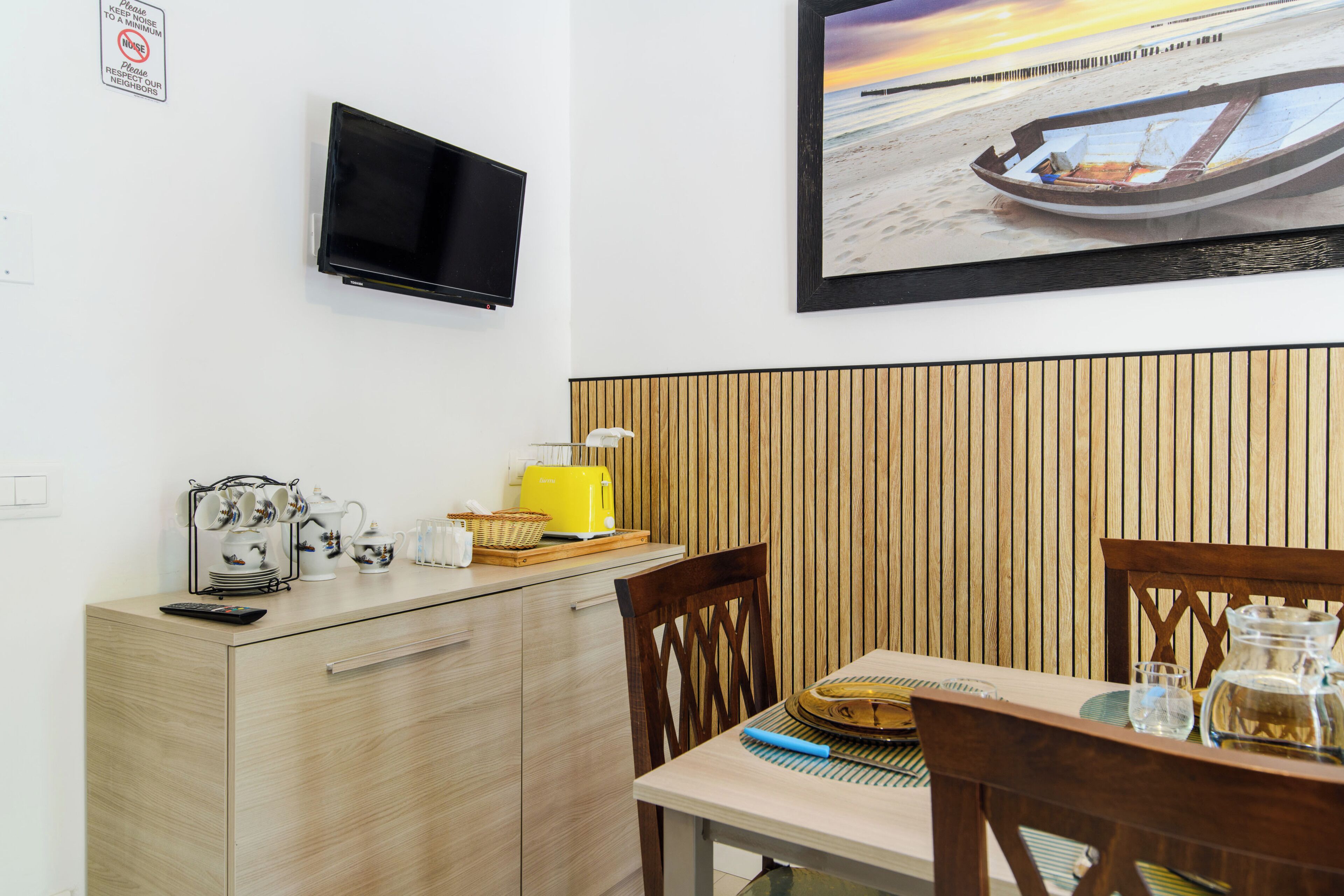 Een kamer met een aan de muur bevestigde televisie, een houten kast, een eettafel met stoelen en een schilderij van een boot aan de muur.
