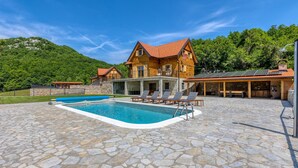 Une maison en bois à deux étages, dotée d’une piscine, d’une terrasse en pierre et offrant une vue sur les montagnes.