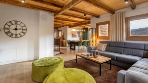 Un salon moderne avec un plafond en bois, une horloge murale ronde, un canapé, des poufs et, au fond, un coin repas.
