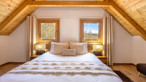 Une chambre avec un plafond haut, des poutres en bois, deux fenêtres avec des rideaux, un lit avec une literie blanche et des lampes de chevet.